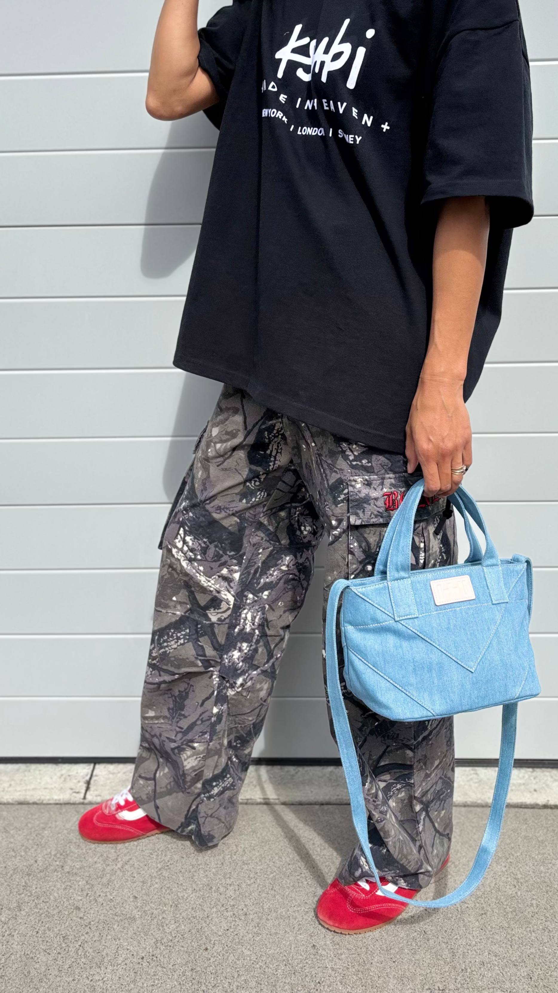 Person wearing a black t-shirt with white text, camouflage pants, and red shoes, holding a blue handbag against a gray wall.